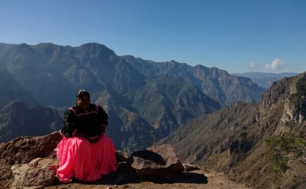 A woman wearing a pink skirt and black clothes sits with a valley in the background.
