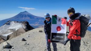 高山をバックにサングラスをした男女二人が山の頂上で日本とメキシコの国旗を掲げている