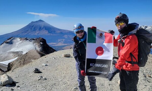 A man and a woman wearing sunglasses with high mountains in the background hold up the national flags of Japan and Mexico on the top of the mountain