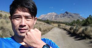 青いシャツを着た男性が険しい山を背景にガッツポーズをしている