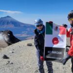 高山をバックにサングラスをした男女二人が山の頂上で日本とメキシコの国旗を掲げている